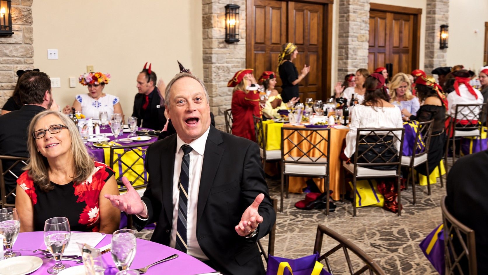 A Galaween guest in a suit reacting with open arms at his table, with costumed guests behind him